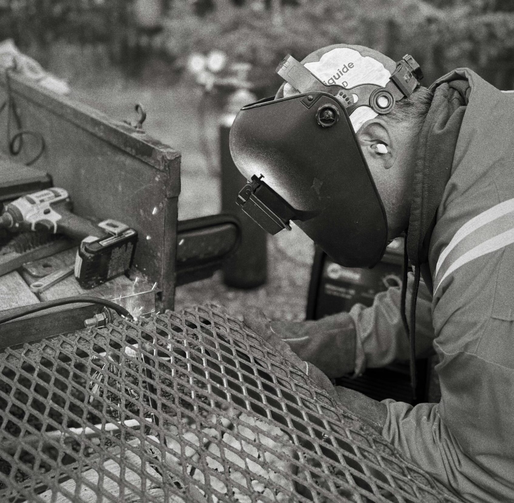 A welder wearing a protective helmet works on metal outdoors.